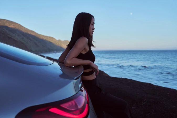 Asian woman in black leans against silver car by ocean at dusk, with mountains in background under a blue sky with the moon.