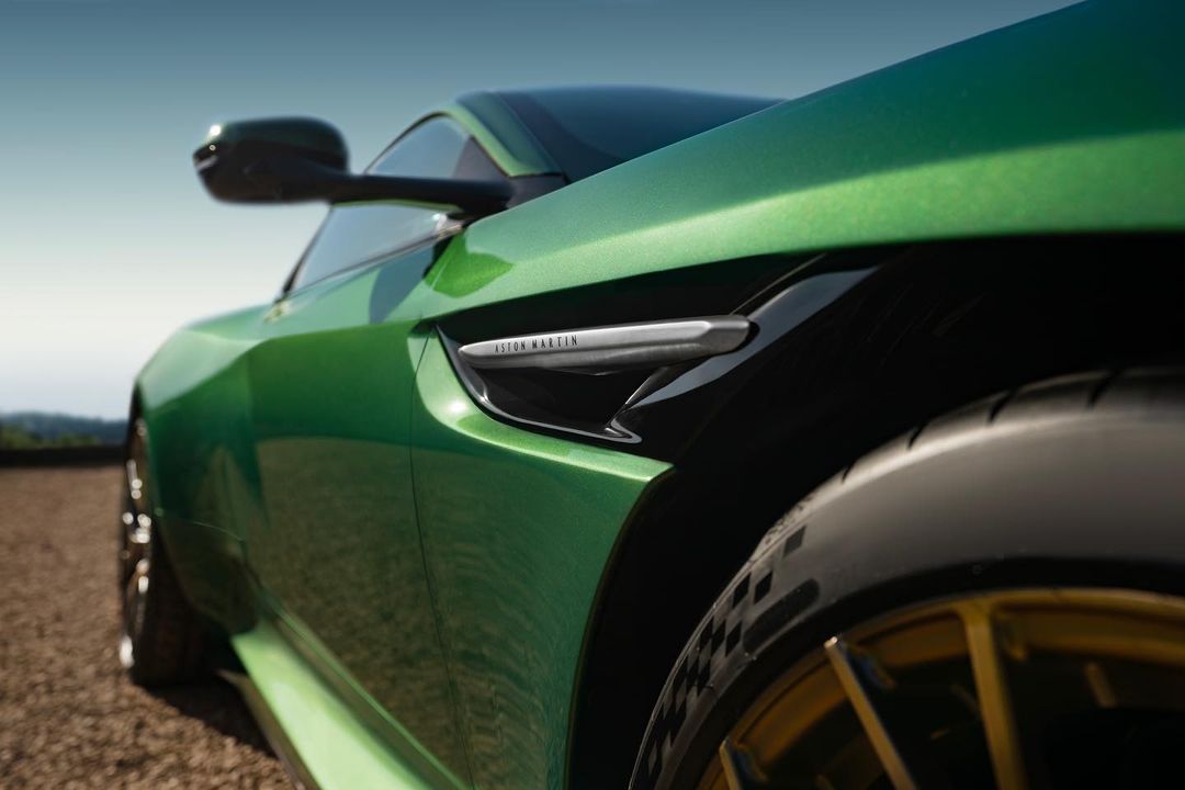 Close-up of a sleek, green Aston Martin showcasing its sporty design, glossy finish, and gold rims against a soft outdoor backdrop.