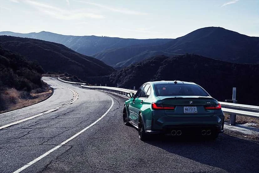 Green BMW M3 parked on a scenic mountain road, with a winding asphalt road and dark, forested mountains in the background under a bright sky.