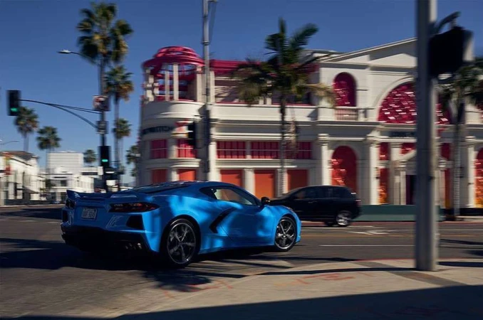 A blue Chevrolet Corvette speeds down a city street on a sunny day with palm trees and white buildings in the background.