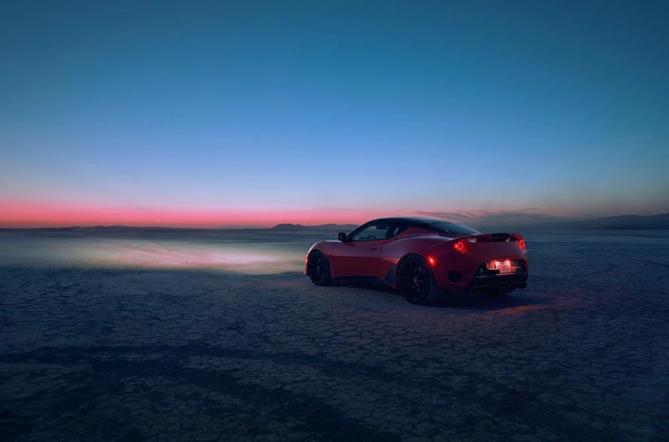 Red sports car on a cracked dry lakebed at dusk, featuring clear blue sky, pink horizon, and faint mountains in the distance.