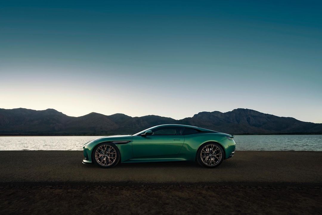 Side view of a green Aston Martin coupe parked on a gray asphalt road near a lake with mountains and a blue and white sky in the background.