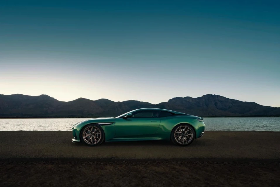 Side view of a green Aston Martin coupe parked on a gray asphalt road near a lake with mountains and a blue and white sky in the background.