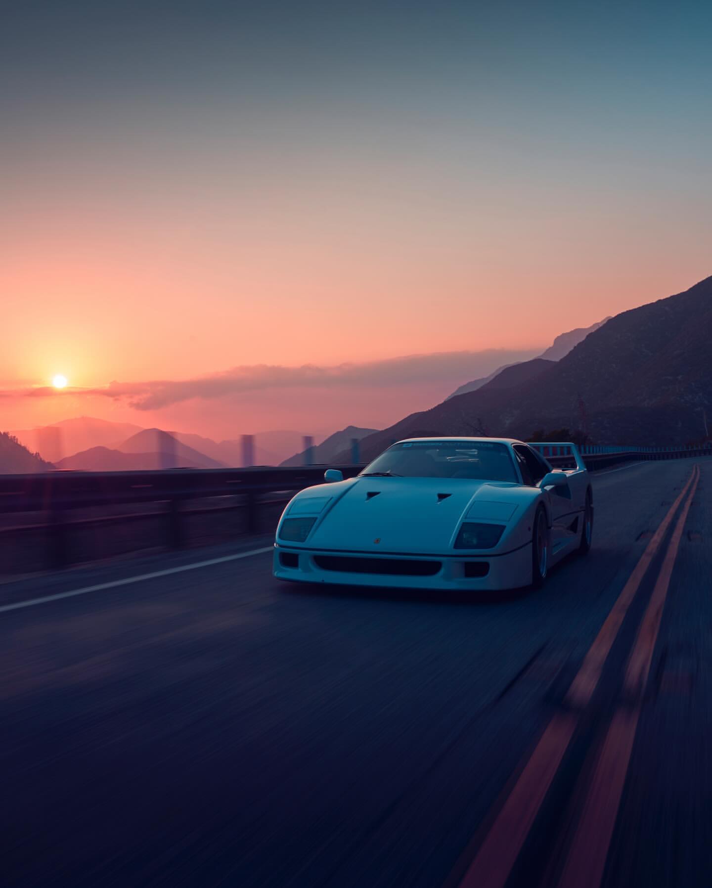 White Ferrari F40 driving on a winding road at sunset with mountain views, capturing an automotive lifestyle aesthetic.