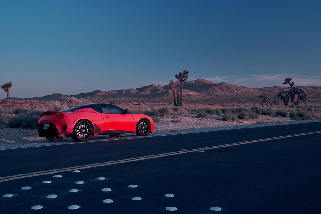 Red sports car parked on the side of a road in a desert landscape with mountains and Joshua trees under a blue sky at dusk.