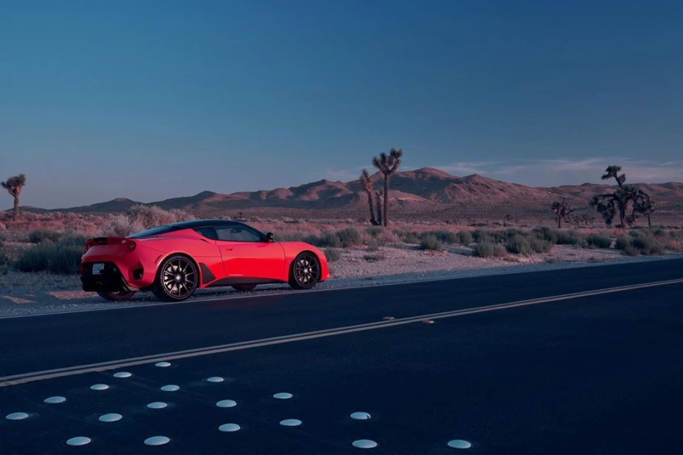 Red sports car parked on the side of a road in a desert landscape with mountains and Joshua trees under a blue sky at dusk.