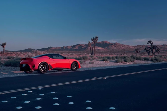 Red sports car parked on the side of a road in a desert landscape with mountains and Joshua trees under a blue sky at dusk.