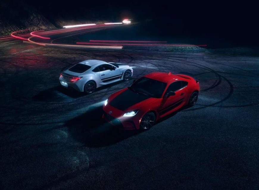 Two-door coupe automobiles, one red and one white, are parked on the asphalt in an outdoor, low-lit automotive presentation.