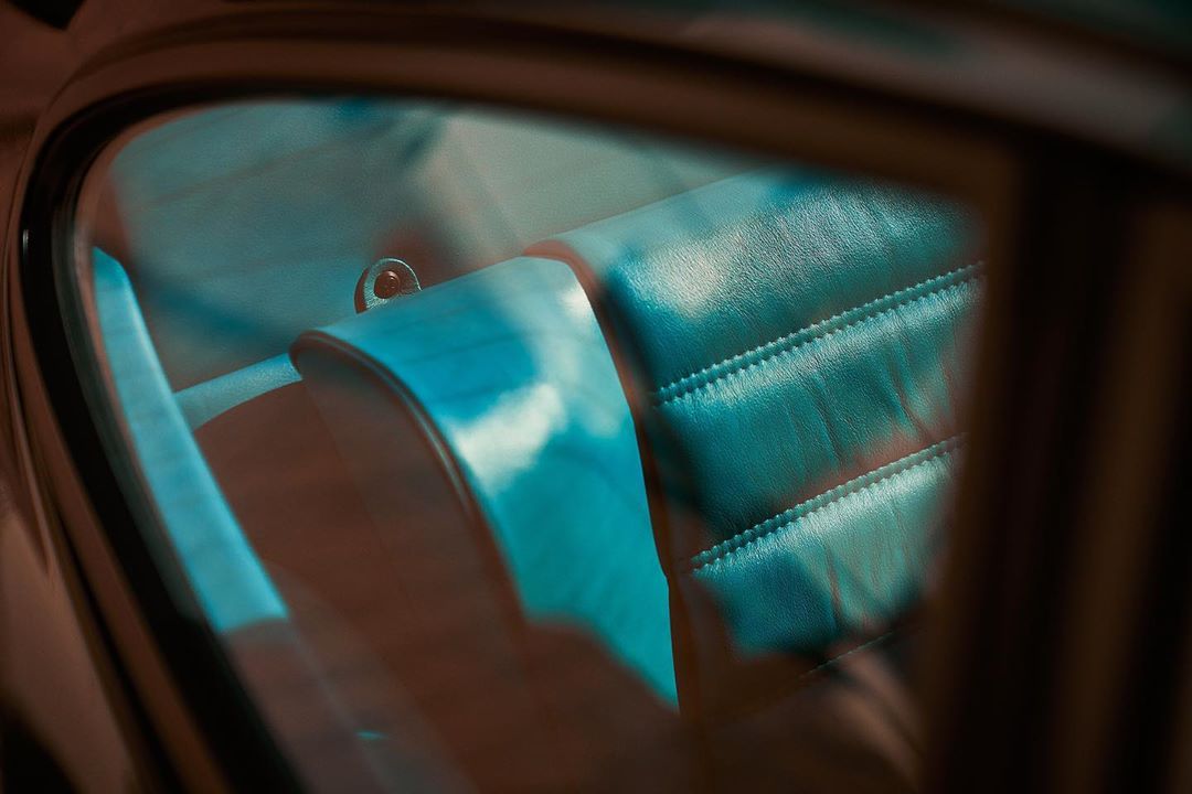 Close-up of an interior of an old car with teal leather seats viewed through the window with visible window frame and blurry reflections.
