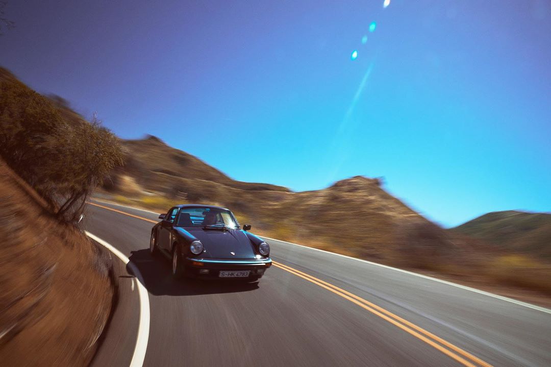 Black classic Porsche 911 Carrera driving on an asphalt road through a mountainous landscape under a clear blue sky on a sunny day.