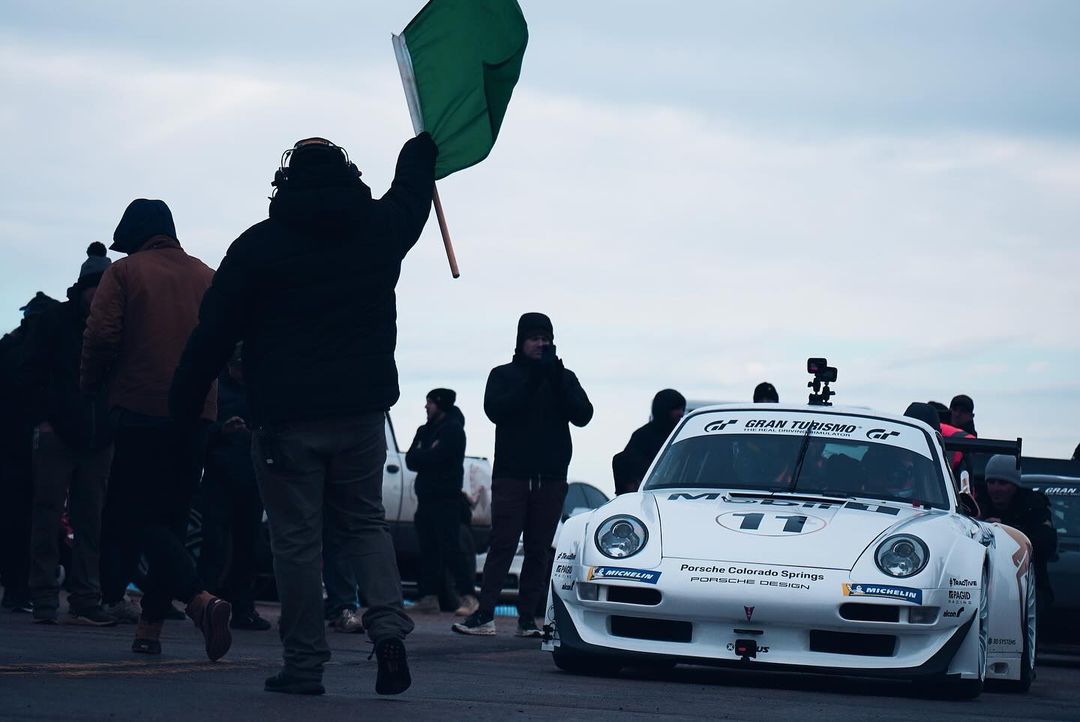 White Porsche race car on track with officials and spectators in overcast conditions, green flag visible in foreground. Automotive photography.