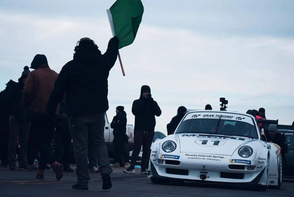 White Porsche race car on track with officials and spectators in overcast conditions, green flag visible in foreground. Automotive photography.