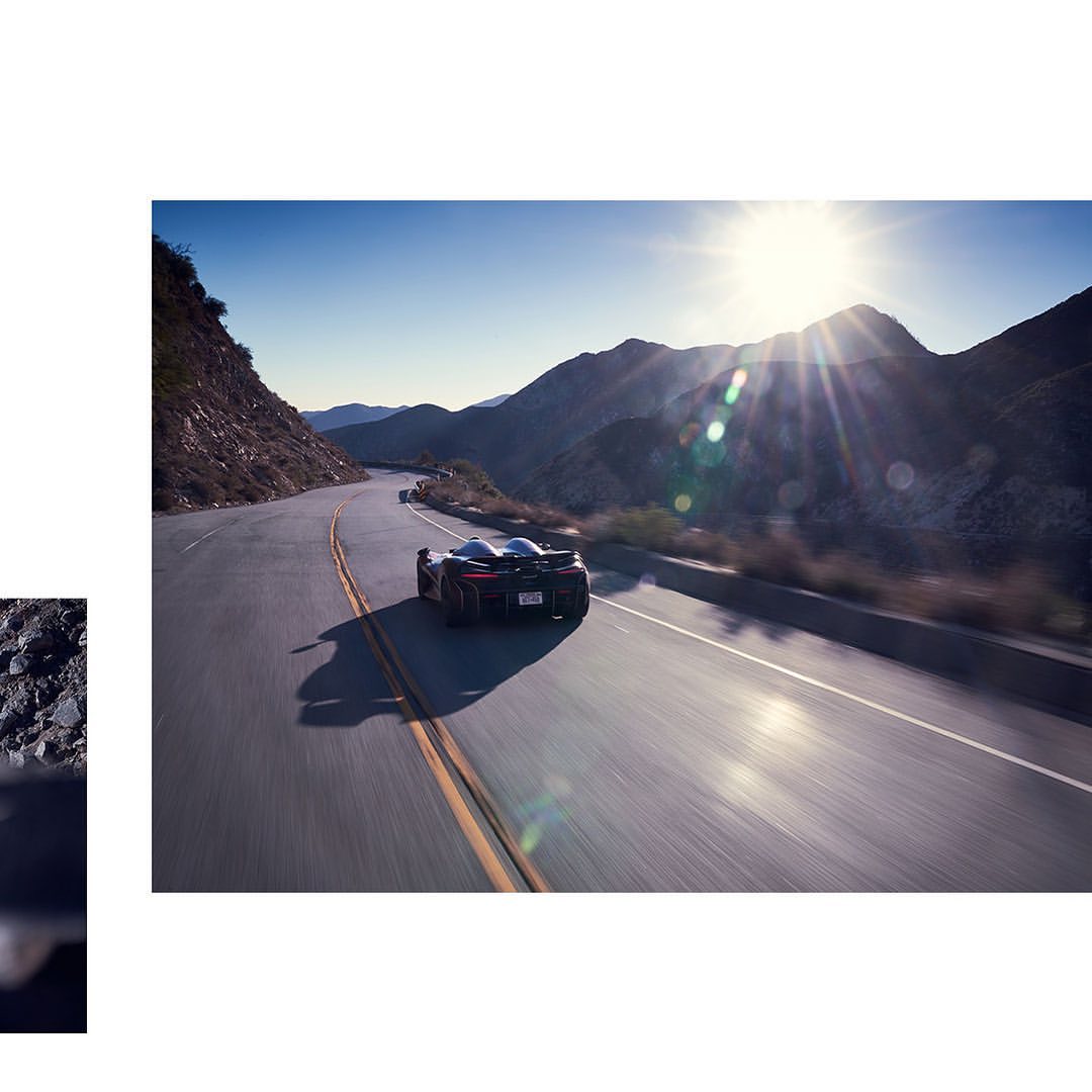 Rear view of a sleek, dark sports car speeding along a winding asphalt road through a mountainous landscape on a sunny day.