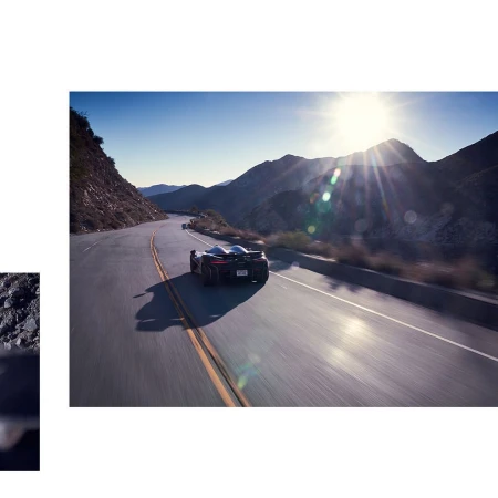 Rear view of a sleek, dark sports car speeding along a winding asphalt road through a mountainous landscape on a sunny day.