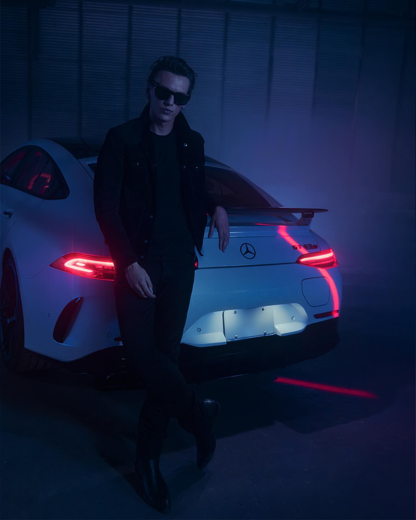 An adult male leans against a white Mercedes AMG GT, wearing a dark jacket and sunglasses, set against a dark, blue-lit background.