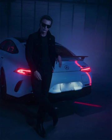 An adult male leans against a white Mercedes AMG GT, wearing a dark jacket and sunglasses, set against a dark, blue-lit background.