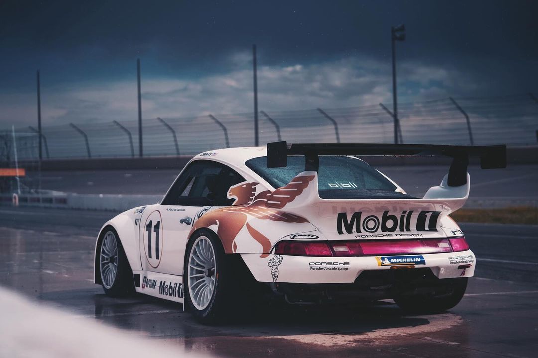 White Porsche race car with Mobil logo on the side, number 11, parked on a racetrack. Rear angled view of the vehicle.