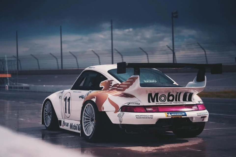 White Porsche race car with Mobil logo on the side, number 11, parked on a racetrack. Rear angled view of the vehicle.