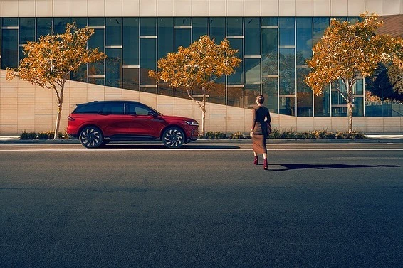 A woman walks away from a red SUV parked on a city street with golden trees and a building with large windows in the background.
