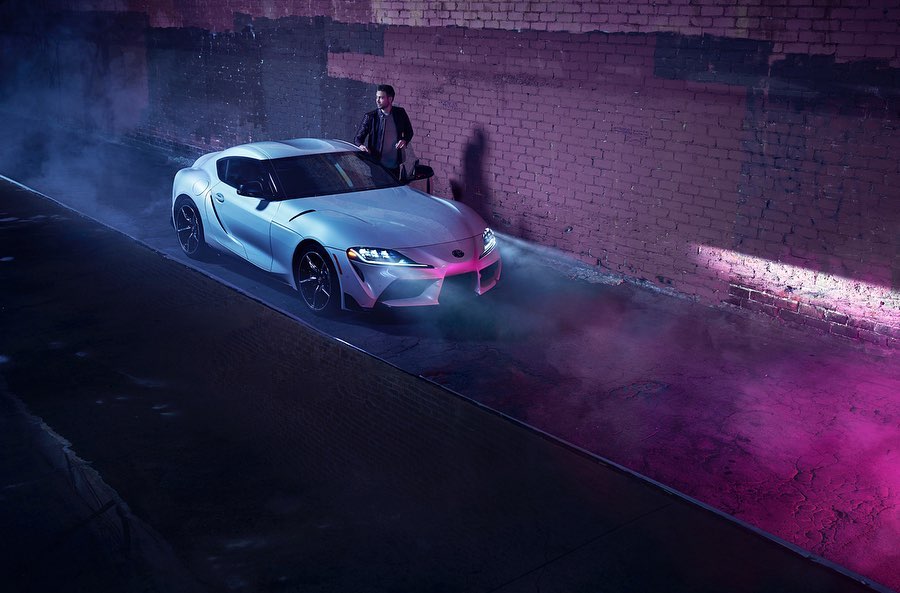 Silver sports car parked on dark street, man standing behind the hood in a jacket and dress pants at night. Automotive and urban theme.