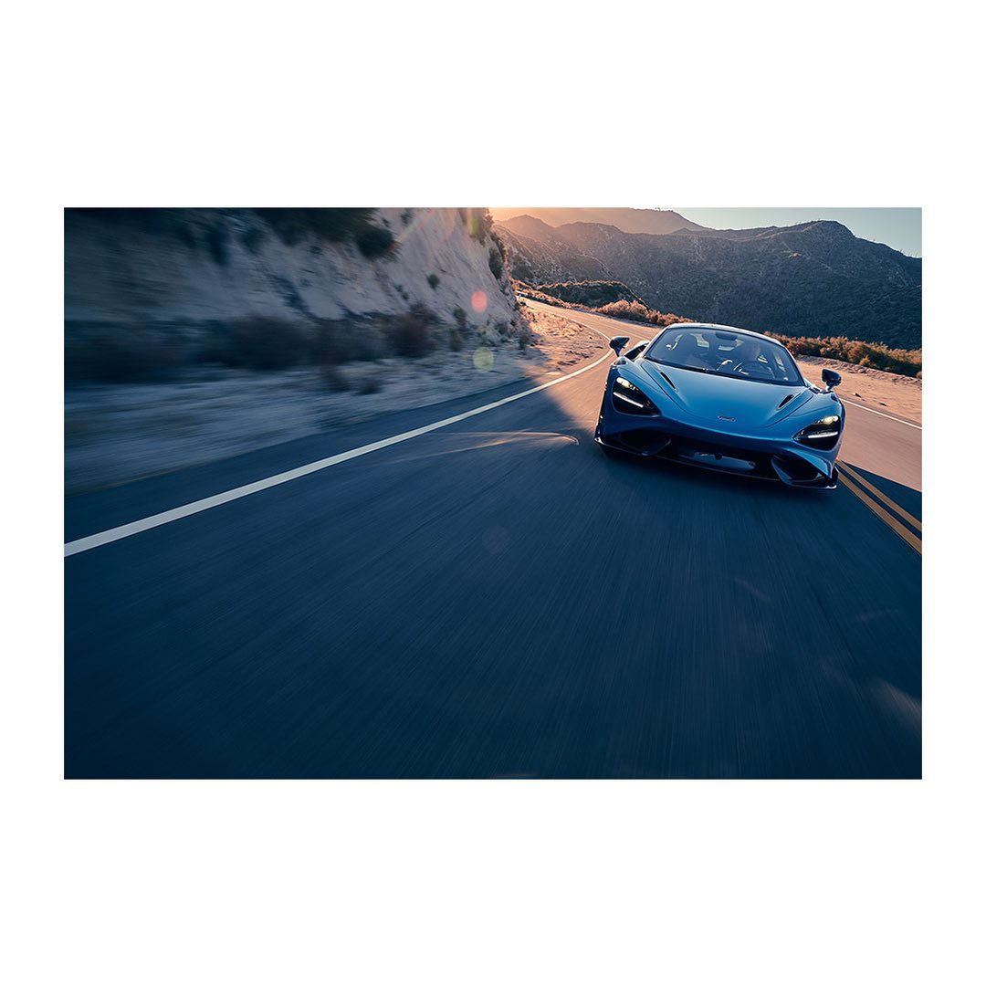 Blue sports car driving on a paved road with a mountain range, blue sky, and the warm glow of sunlight in the background.