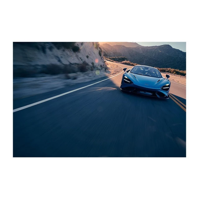 Blue sports car driving on a paved road with a mountain range, blue sky, and the warm glow of sunlight in the background.