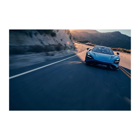 Blue sports car driving on a paved road with a mountain range, blue sky, and the warm glow of sunlight in the background.