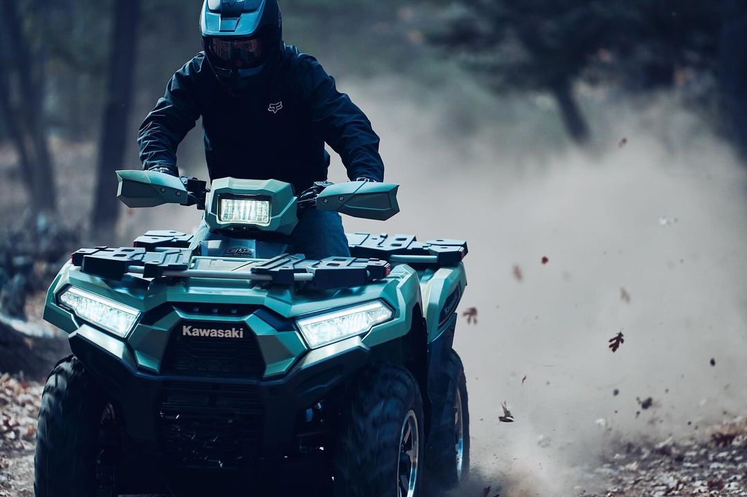 Person riding a green Kawasaki ATV on a dirt path through a wooded area, wearing a helmet and dark clothing. Sport and recreation vehicle.