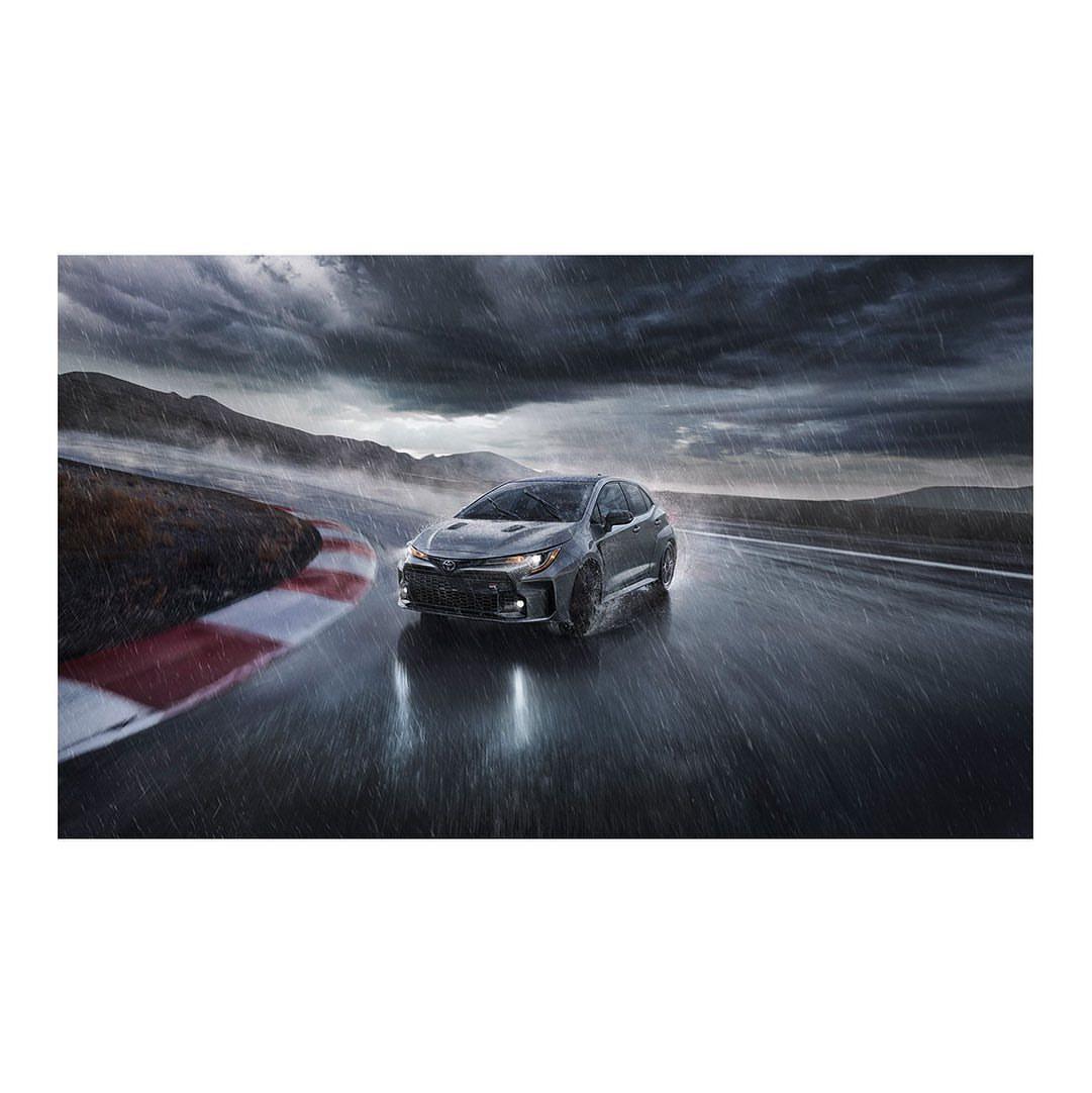 Grey Toyota GR Corolla hatchback driving on a wet racetrack in the rain with moody sky. Automotive product photography.