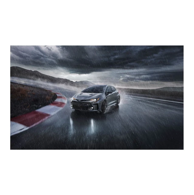 Grey Toyota GR Corolla hatchback driving on a wet racetrack in the rain with moody sky. Automotive product photography.