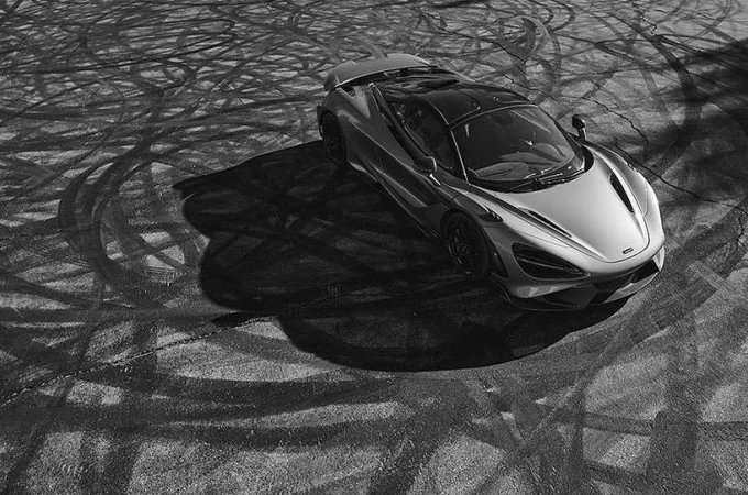 Overhead black and white shot of a silver Mclaren sports car on a cracked asphalt surface marked with circular tire track patterns.