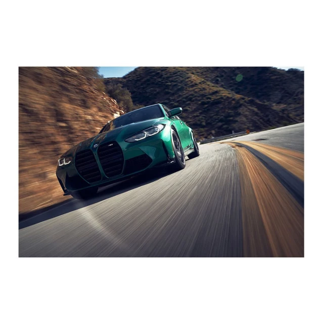 Green BMW M4 speeding along mountain road on a bright day. Panning shot captures motion, showcasing car's design and performance.