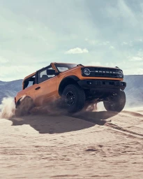 Orange SUV driving across a sandy desert landscape, kicking up dust with bright daylight and mountains in the background