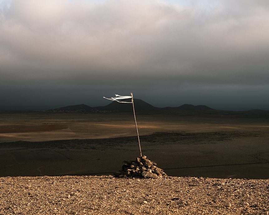 A cairn with a tattered white flag on a wooden pole sits atop a hill in a tan desert landscape under a cloudy sky.