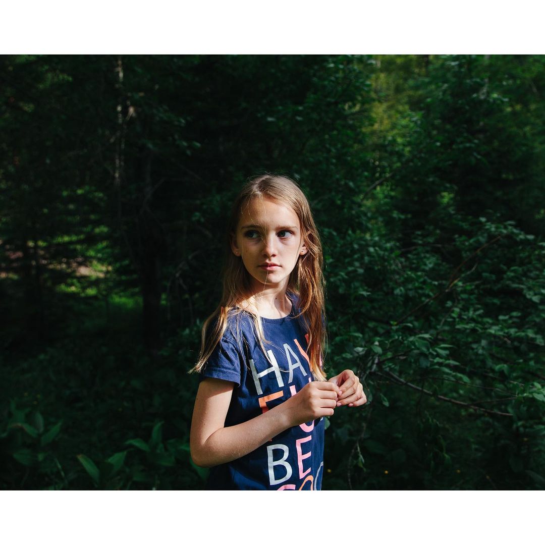 A young girl with blonde hair in a blue t-shirt stands in a lush green forest, looking off to the side in a natural setting.