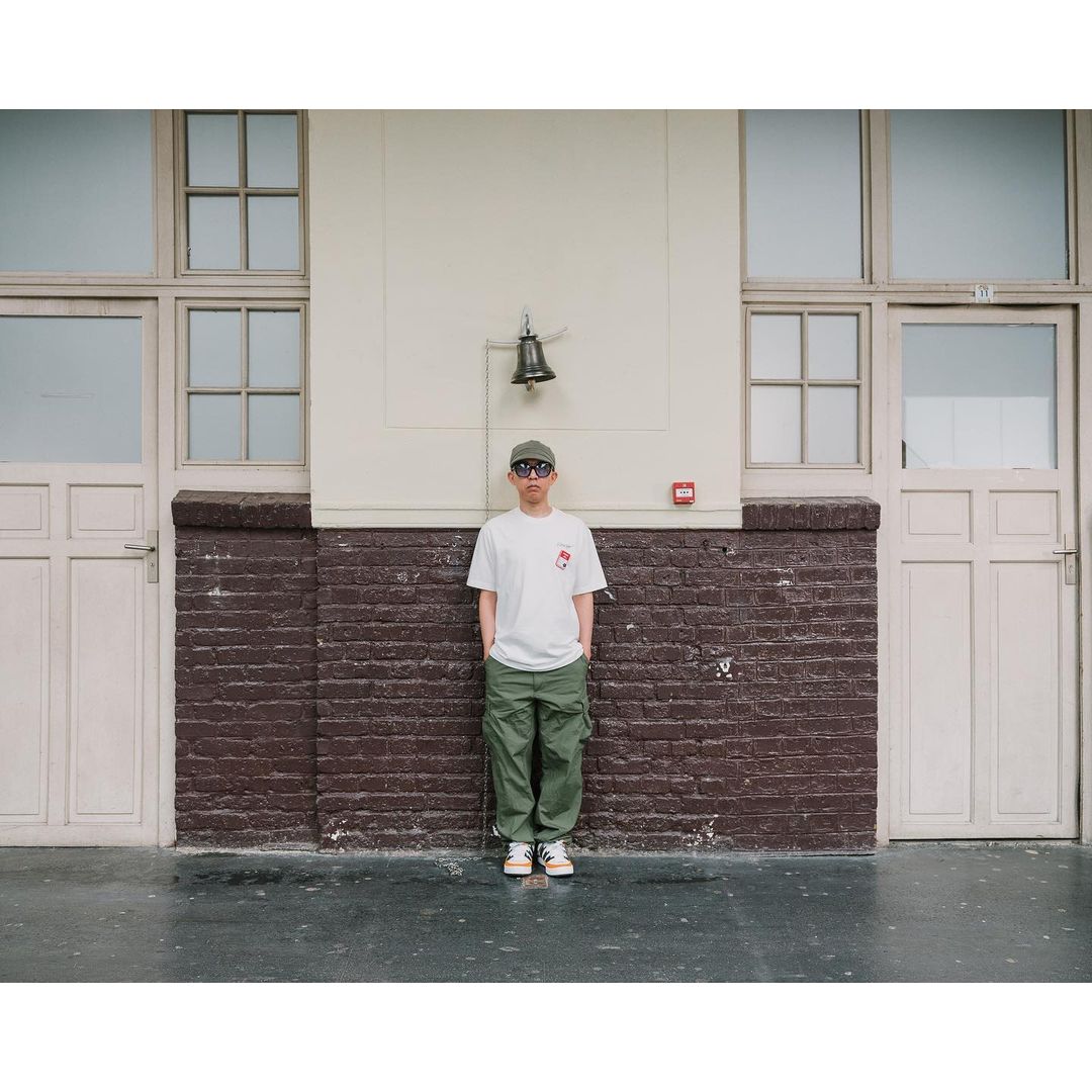 A young person stands outdoors wearing a white t-shirt, green cargo pants, sunglasses and a hat against a textured wall backdrop.