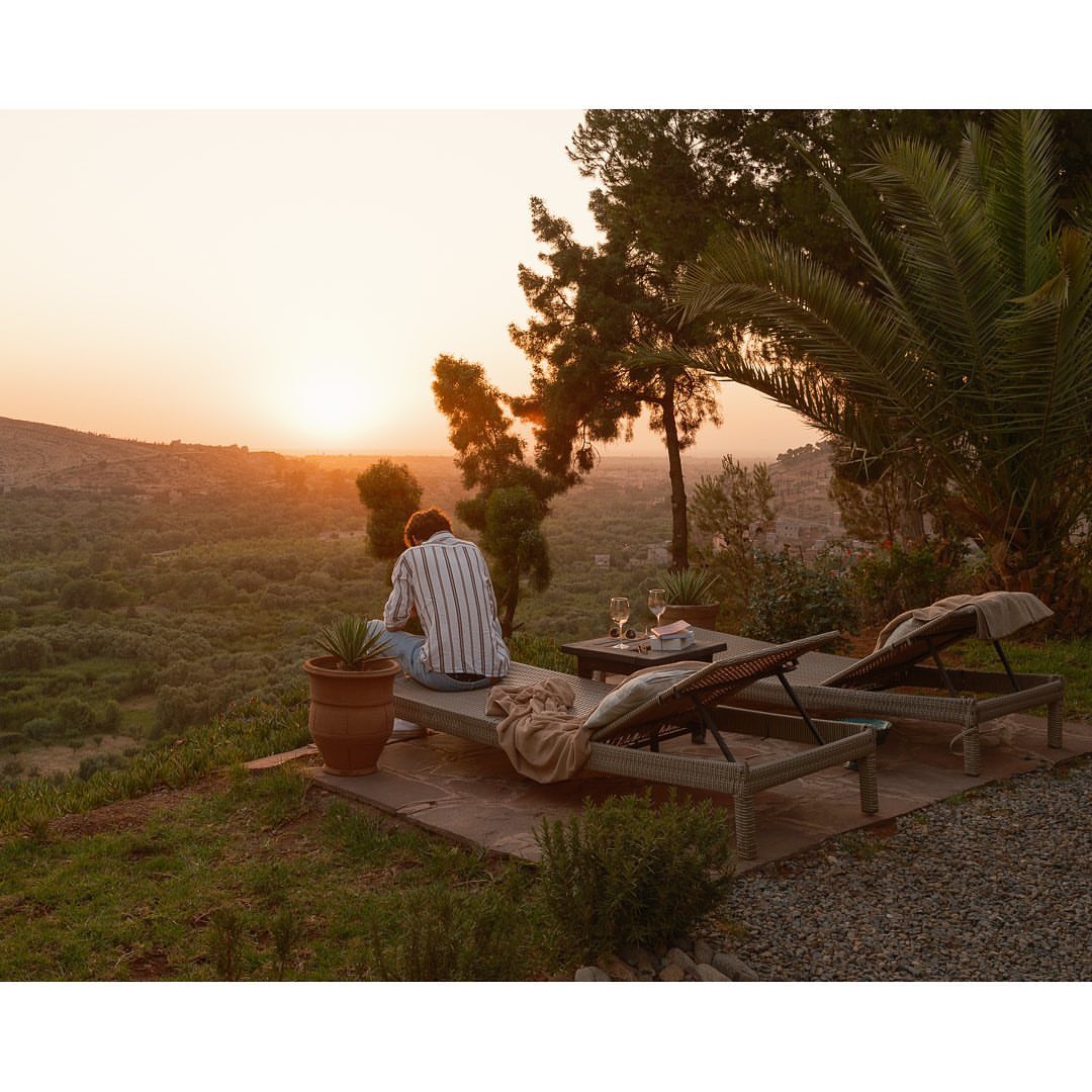 A man sits on a lounge chair outside watching the sunset over a lush valley with wine glasses and a table nearby.