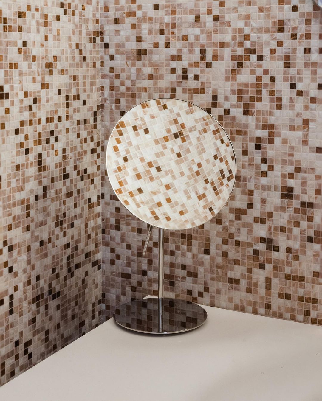 Interior bathroom corner with mosaic tiled walls and a modern round vanity mirror on a white countertop. Warm neutral color palette.