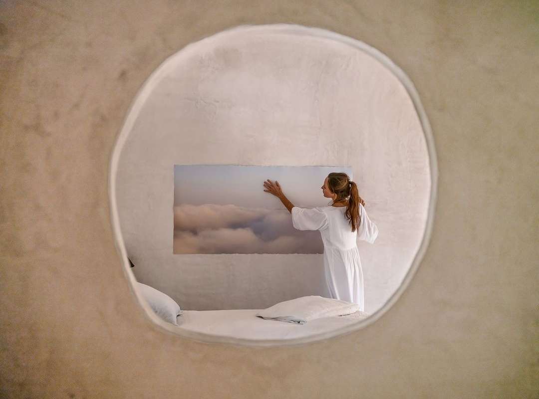 Indoor shot of a woman in a white dress standing near a window looking at an outdoor photograph of clouds in a bedroom.
