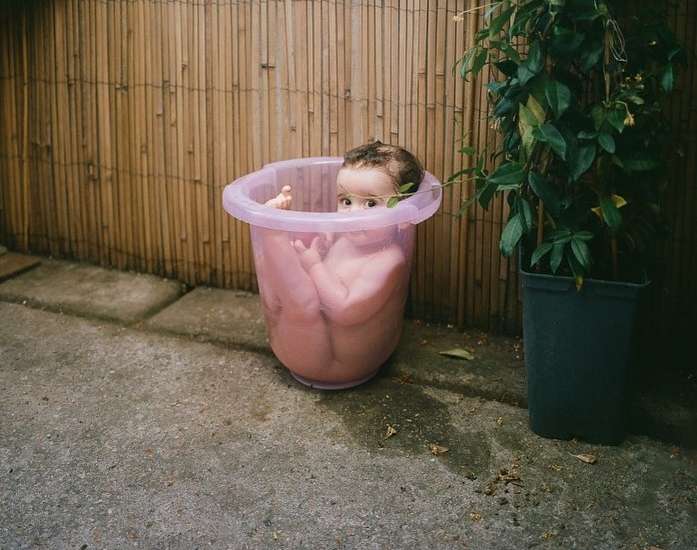 Adorable baby sits inside a pink tub outdoors near a bamboo fence and potted plant, captured with soft, natural lighting.