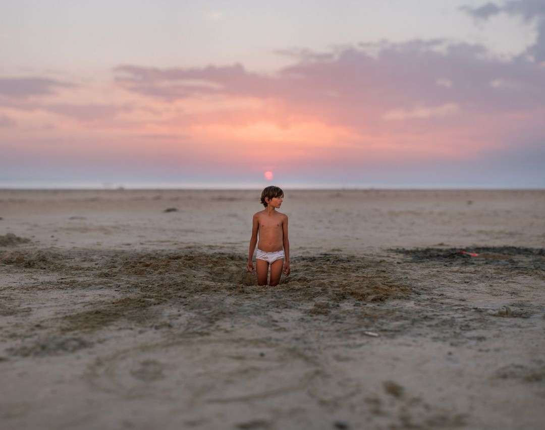 A young child kneels in the sand on a beach at sunset, their gaze directed toward the horizon under a vibrant sky of pink and orange hues.