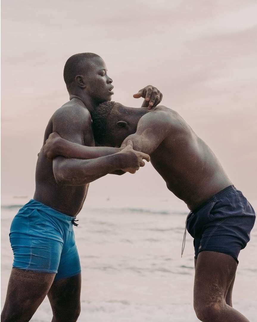 Two dark-skinned men wrestle on a beach with light-colored sand and a light-pink sky in the background, eye-level shot, medium frame.