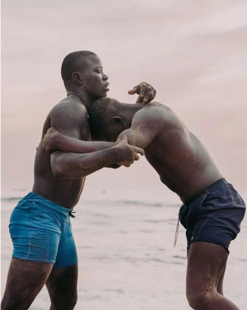 Two dark-skinned men wrestle on a beach with light-colored sand and a light-pink sky in the background, eye-level shot, medium frame.