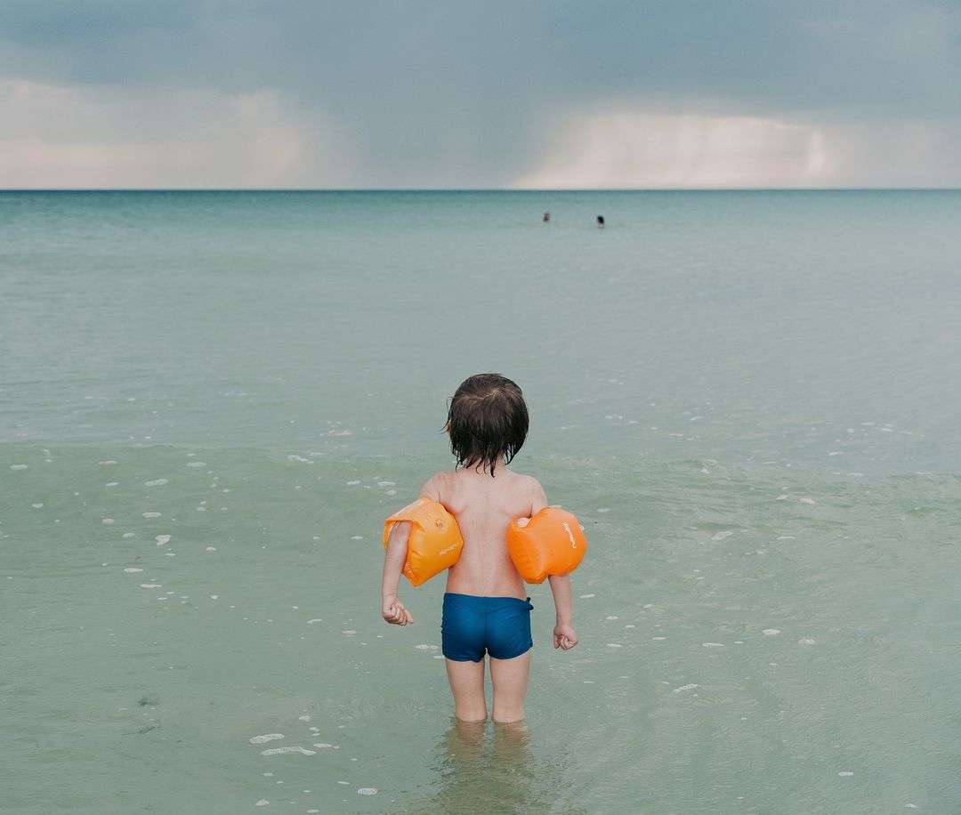 Young child with arm floaties standing in the ocean looking towards the horizon with people visible in the distance on an overcast day.
