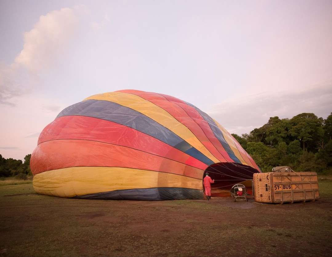 Large deflated hot air balloon in a field at dawn with one person standing by it and a wooden crate in the African landscape.
