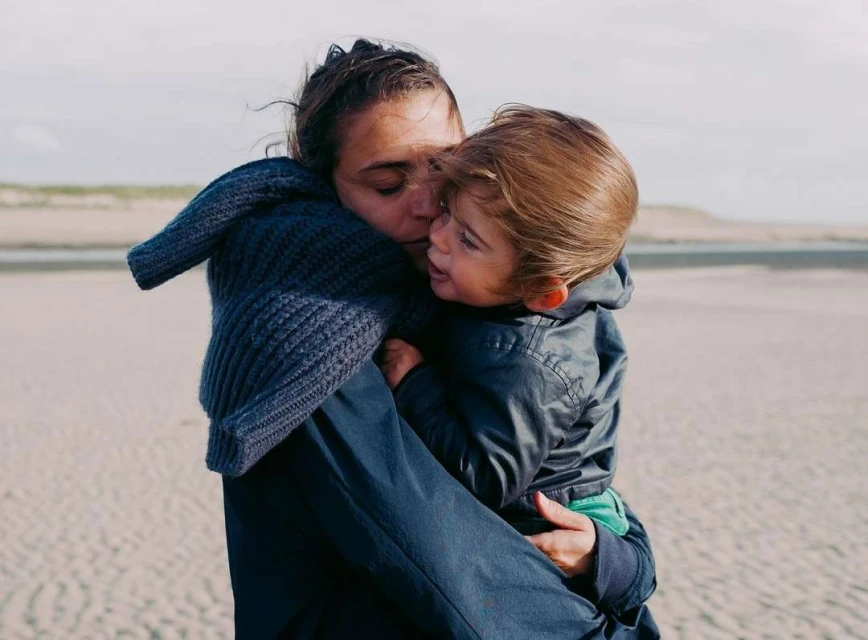 A woman in a blue coat lovingly embraces a toddler on a sandy beach beneath a cloudy sky, capturing a tender family moment.