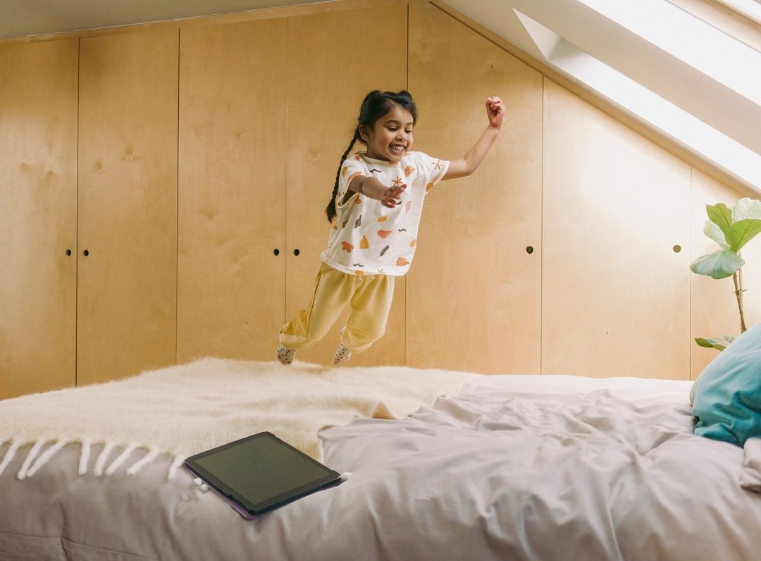 A young child with brown hair jumping on a bed, smiling with arms up in the air, in a brightly lit room with wooden cabinets.