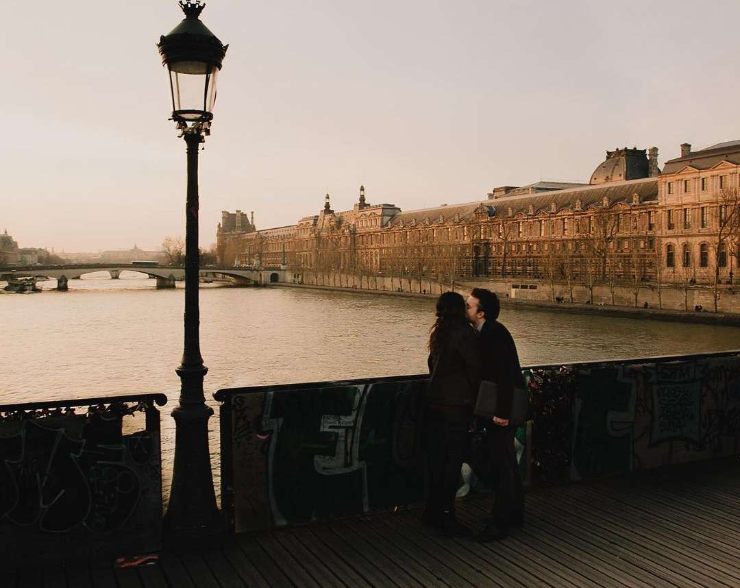 A couple kisses on a bridge in Paris near a vintage lamp post, with the Seine River and historic buildings visible in the background.