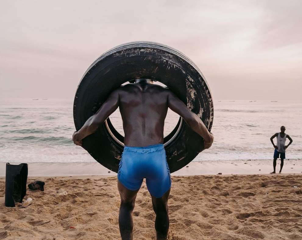African man on beach carrying large tire on his shoulders; another man stands in the background near the water's edge.