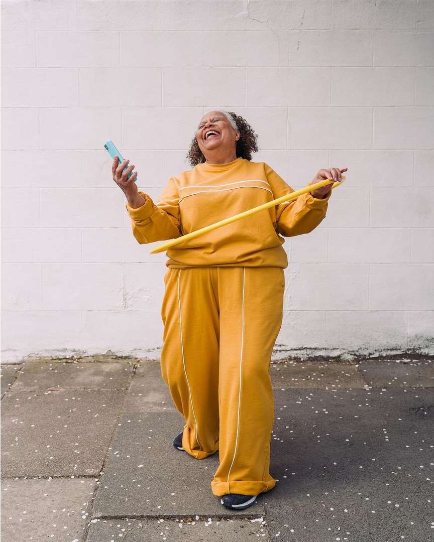 Older woman in yellow tracksuit smiles, holds phone and hula hoop against a white wall and grey pavement in an outdoor setting.